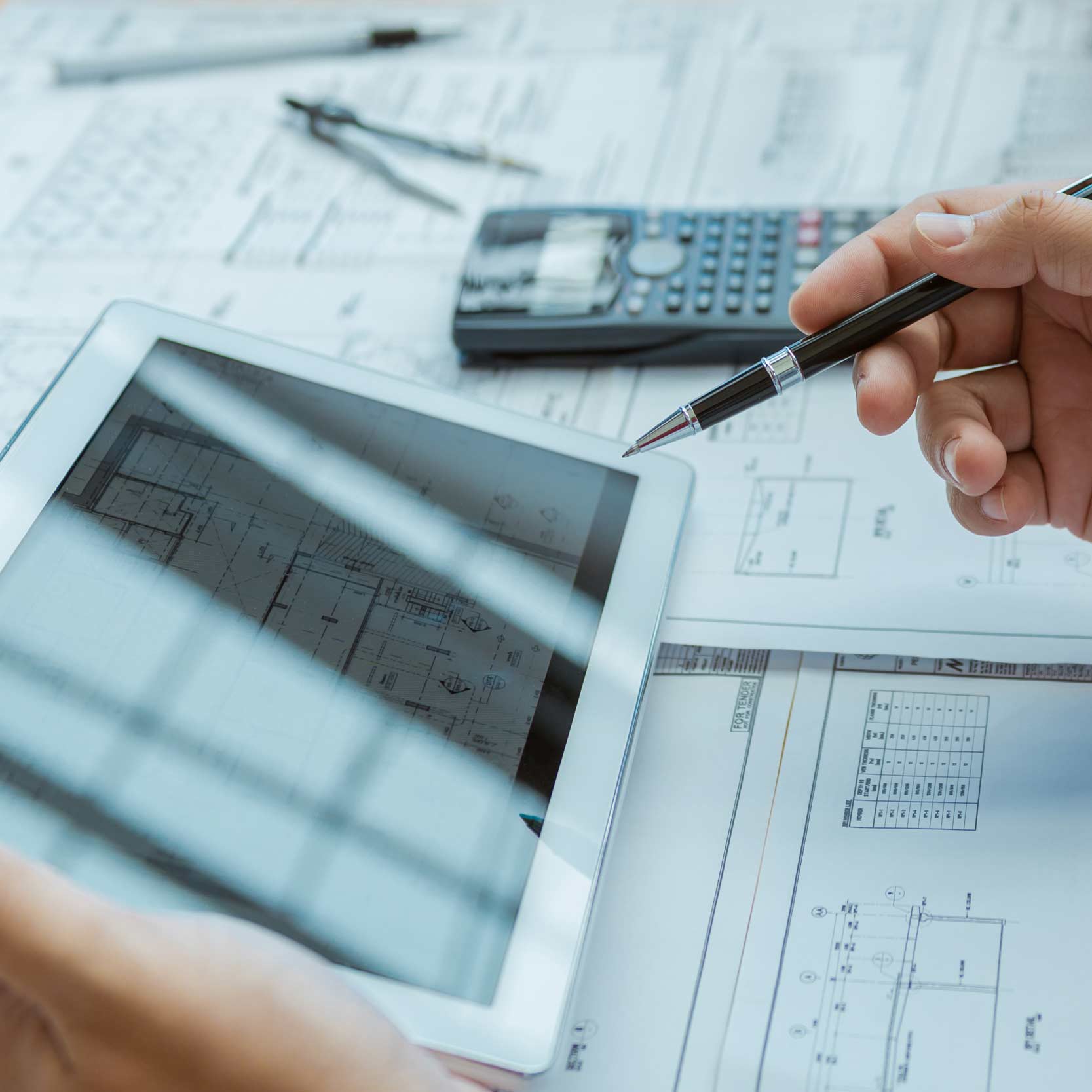 primo piano di una mano che tiene un tablet e l'altra mano che tiene una penna, sullo sfondo calcolatrice, compasso e dei fogli con progetto tecnico 